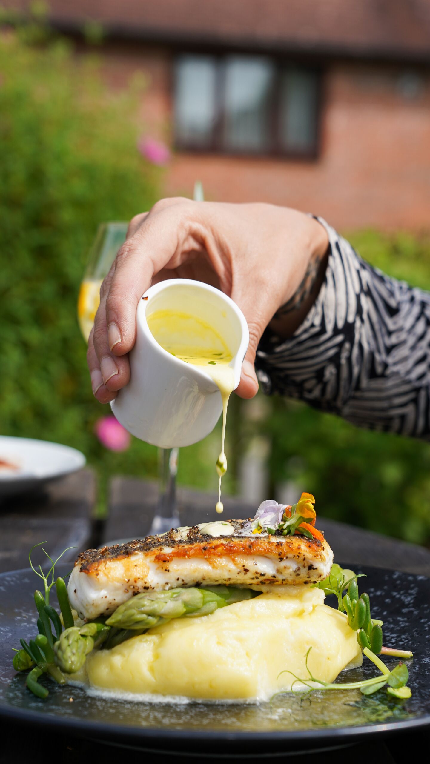 A hand pouring sauce over a fish dish with vegetables and mashed potatoes.