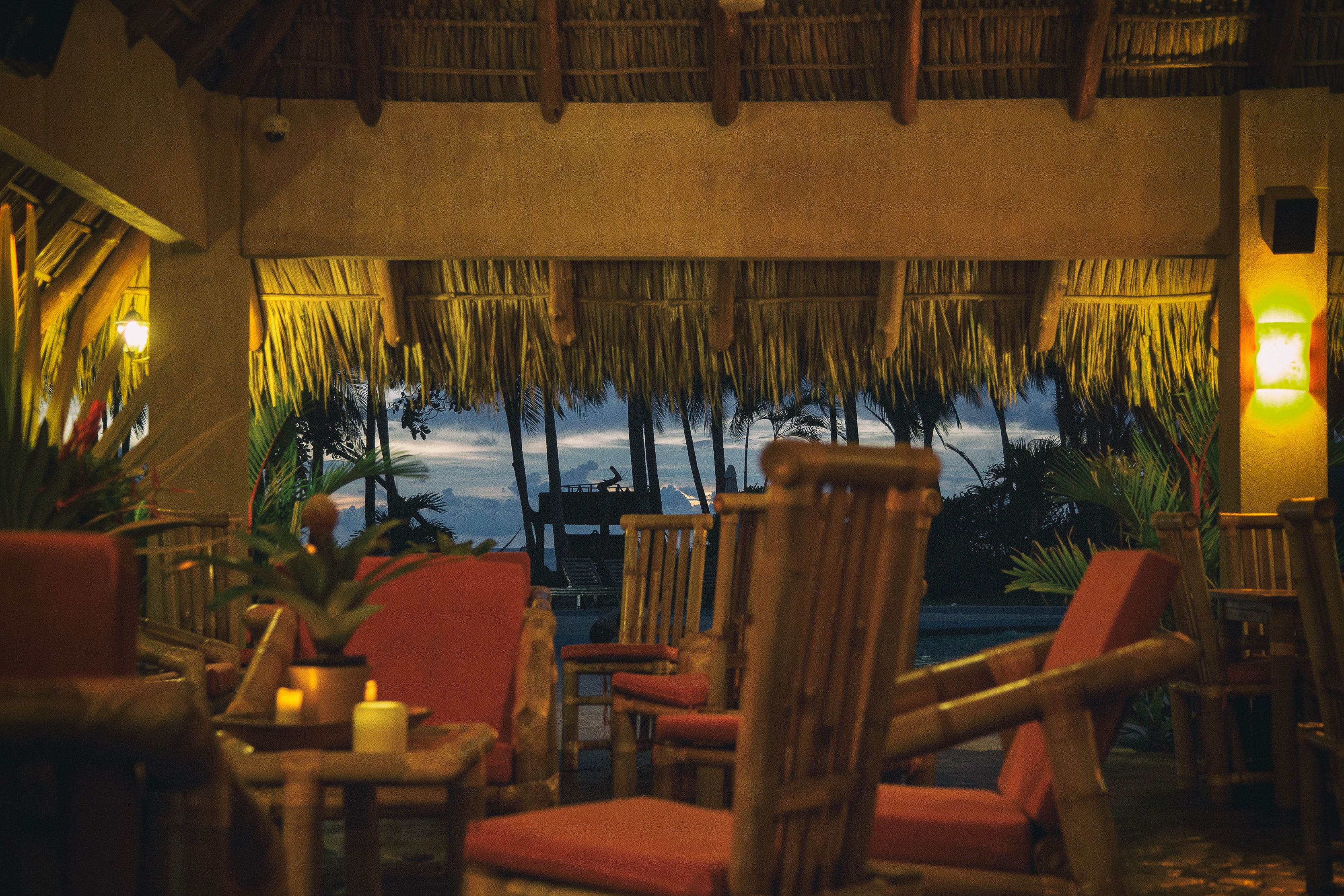 A tropical lounge with thatched roof, wooden furniture, and a view of palm trees and a sunset.