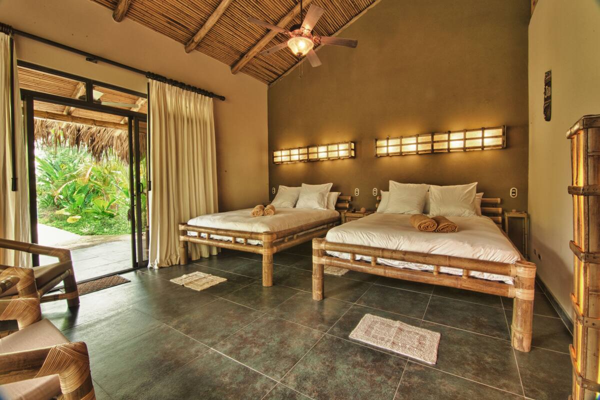 A room with two wooden beds, a ceiling fan, and a view of greenery through a glass door.