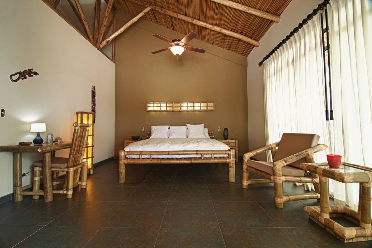 A room with a bed, two chairs, a table, a ceiling fan, and a window with curtains.