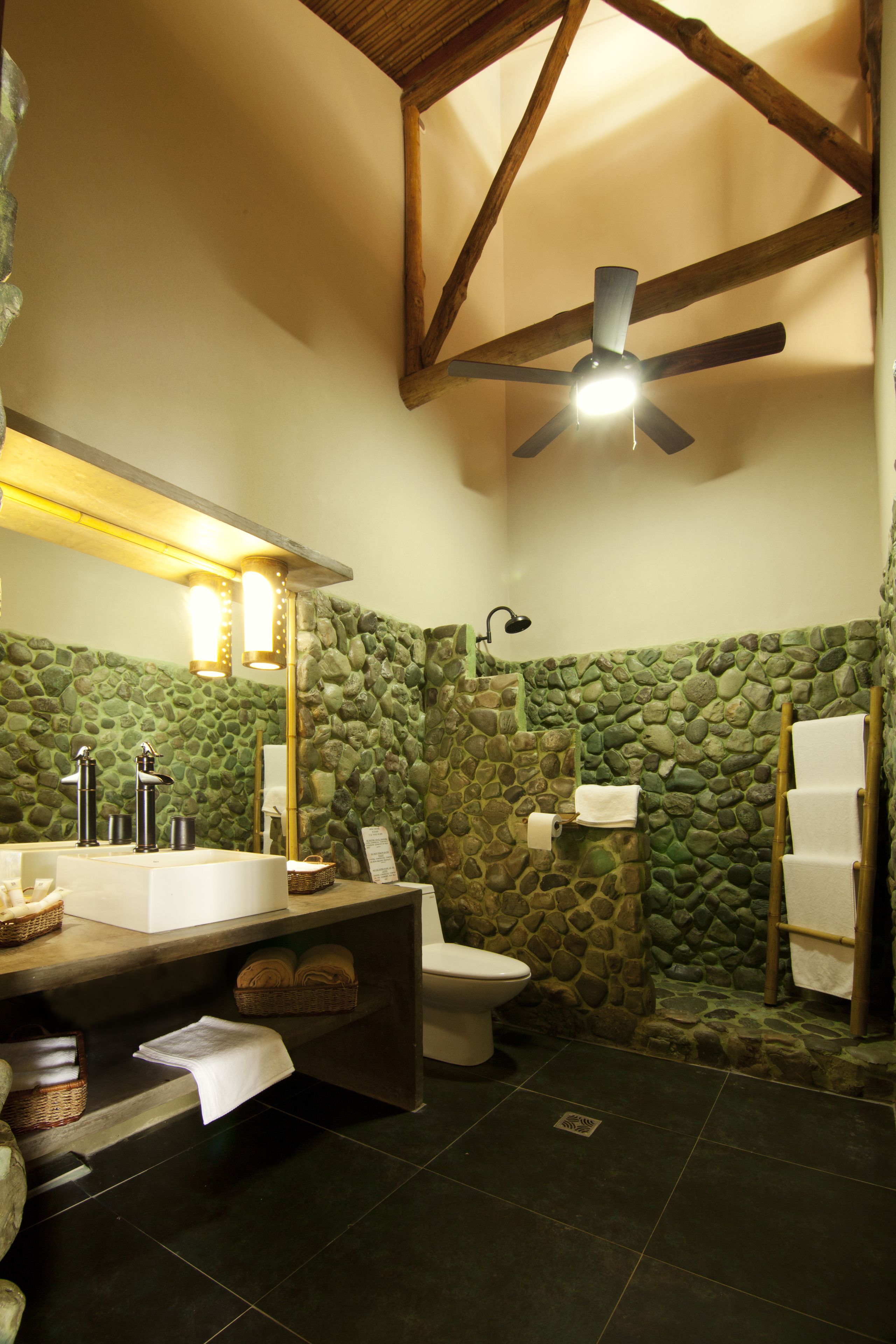 A bathroom with a stone wall, a wooden ceiling, a ceiling fan, a sink, a toilet, and a towel rack.