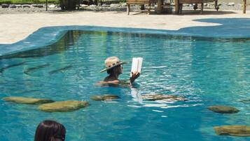 A pool area with a person reading a book, surrounded by palm trees and lounge chairs.