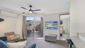 A modern living room with a sofa, a small table, a ceiling fan, and a TV on the wall. - Nelson Towers Motel & Apartments (Nelson Bay)