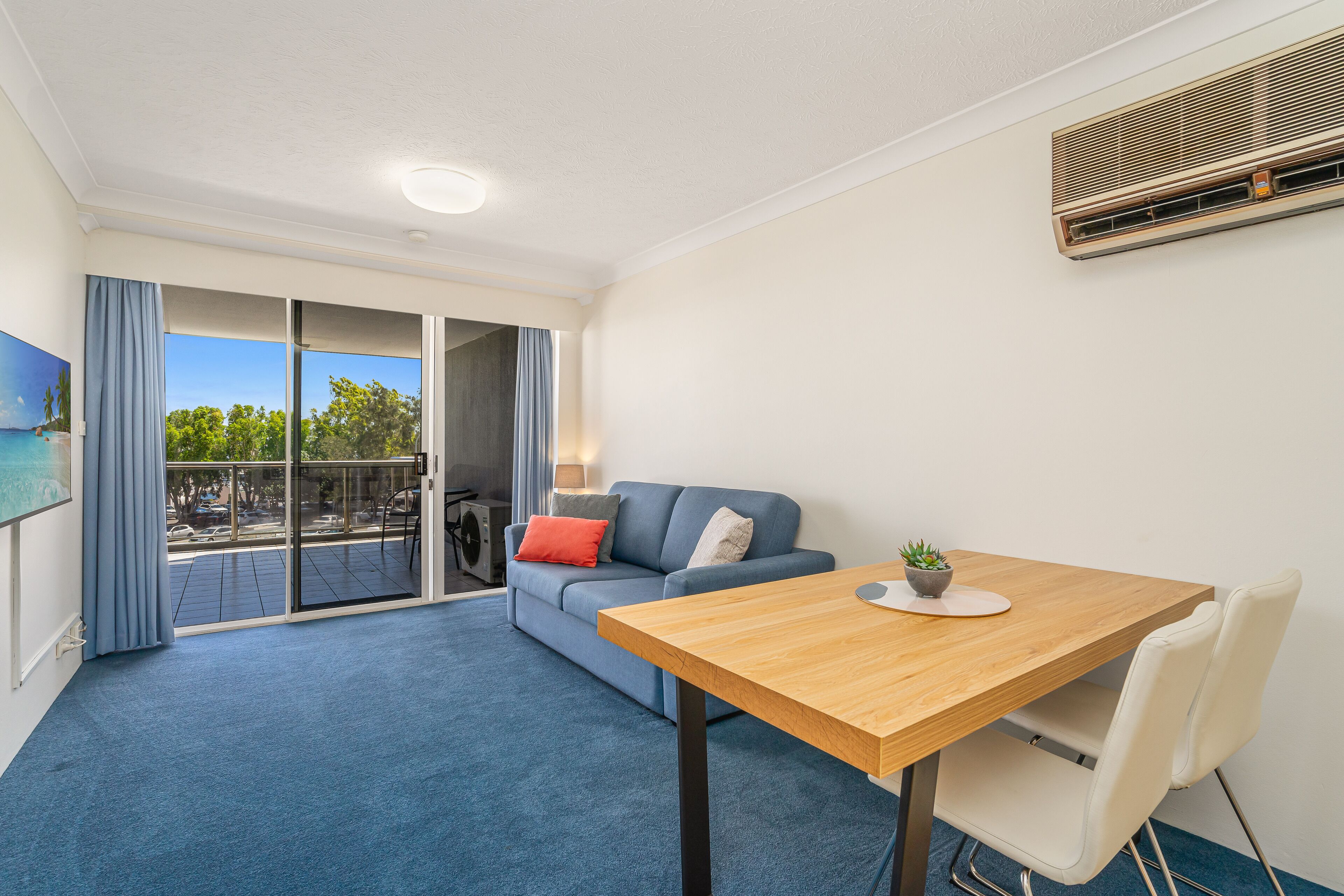 A modern living room with a blue carpet, a wooden table, a sofa with cushions, and a sliding glass door leading to an outdoor area.
