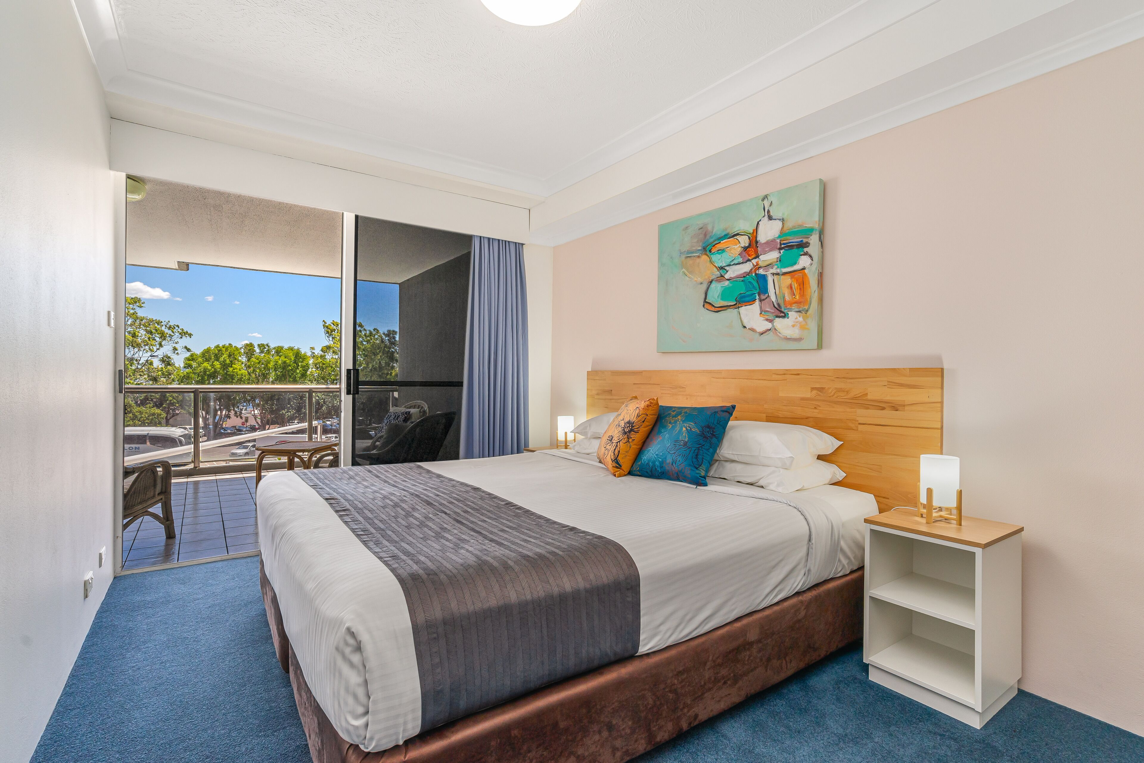A hotel room with a large bed, a wooden headboard, a nightstand, a painting on the wall, and a balcony with a view of the street.