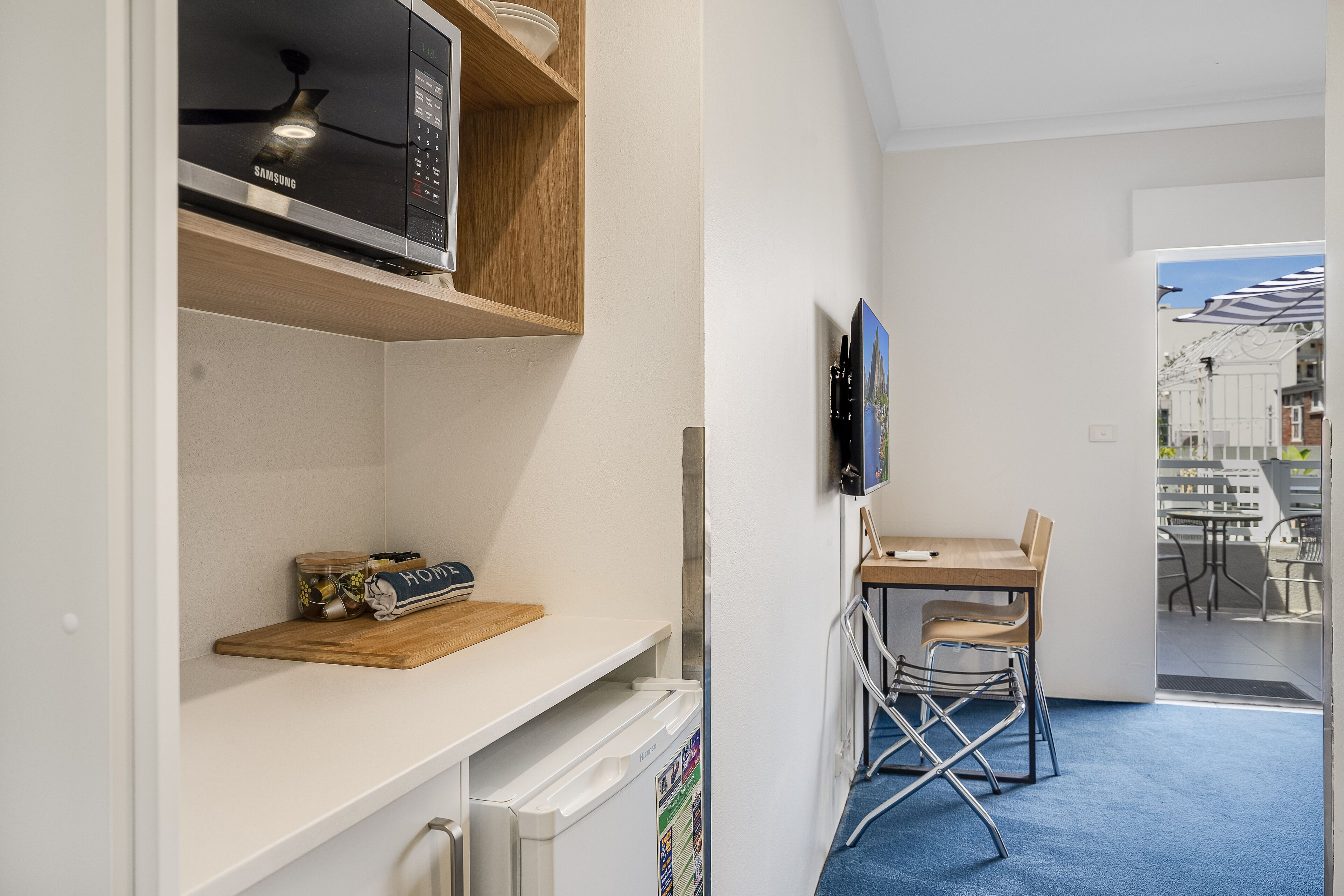 Une cuisine compacte équipée d’un four à micro-ondes, d’une étagère en bois et d’un petit réfrigérateur. On y trouve également un coin repas avec une table et des chaises, et une vue sur l’extérieur grâce à une porte vitrée.