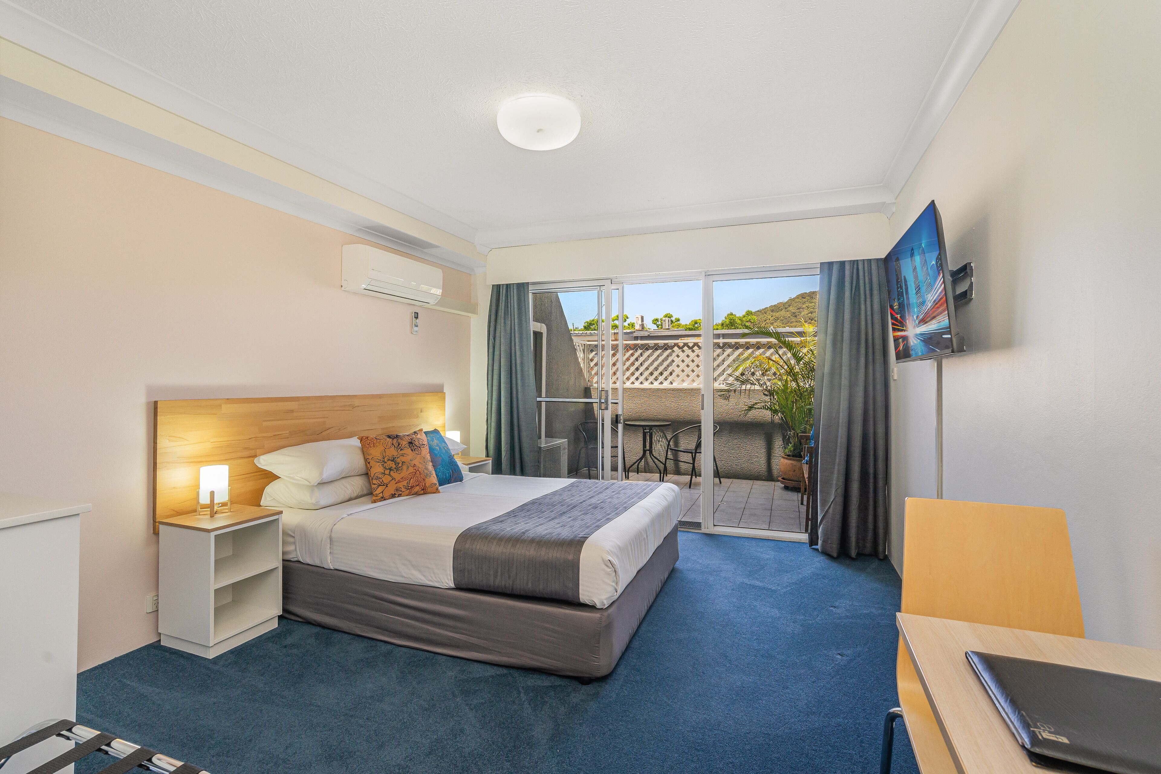 A hotel room with a bed, a desk, a chair, a TV, and a balcony with a view of the outdoors.
