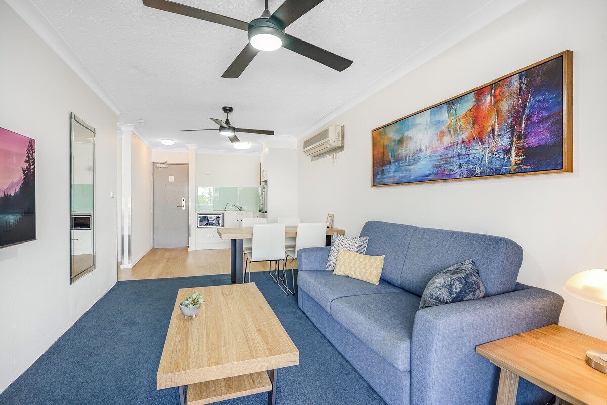 Uma sala de estar moderna com um sofá azul, uma mesa de centro de madeira e um ventilador de teto. Um grande quadro abstrato está na parede, e há uma área de cozinha ao fundo.