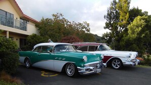 Twee oldtimers op een oprit, met een gebouw en bomen op de achtergrond.