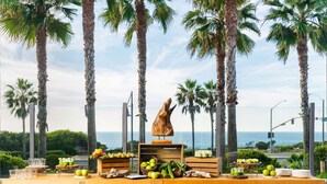 A buffet table with fruits and vegetables, set against a backdrop of palm trees and a view of the ocean. - Cape Rey Carlsbad Beach, a Hilton Resort & Spa (Carlsbad)