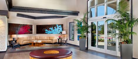 A modern lobby with a beige sofa, a wooden coffee table, and a red circular bench. There are large windows and potted plants.