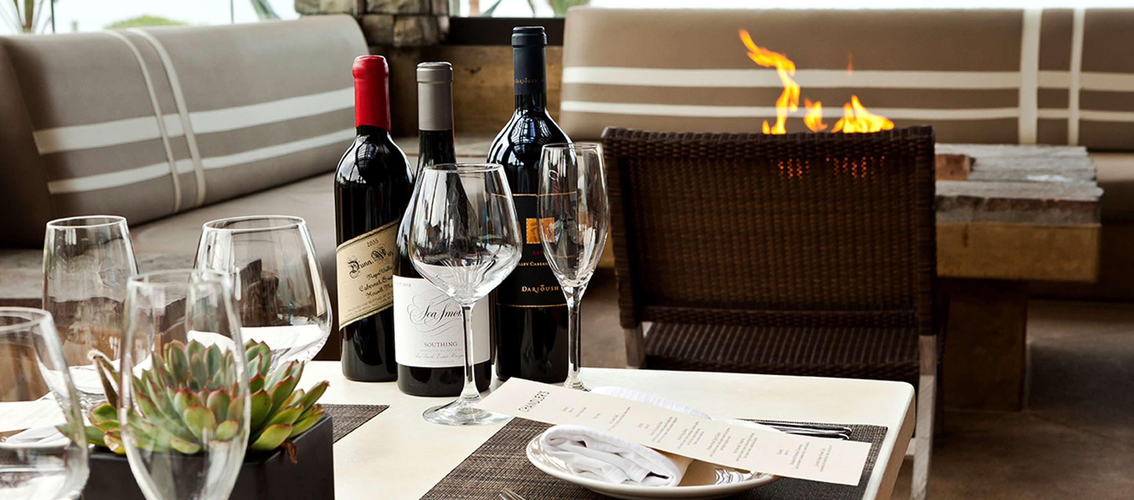 A dining table set with wine bottles, glasses, and a menu, with a fire in the background.