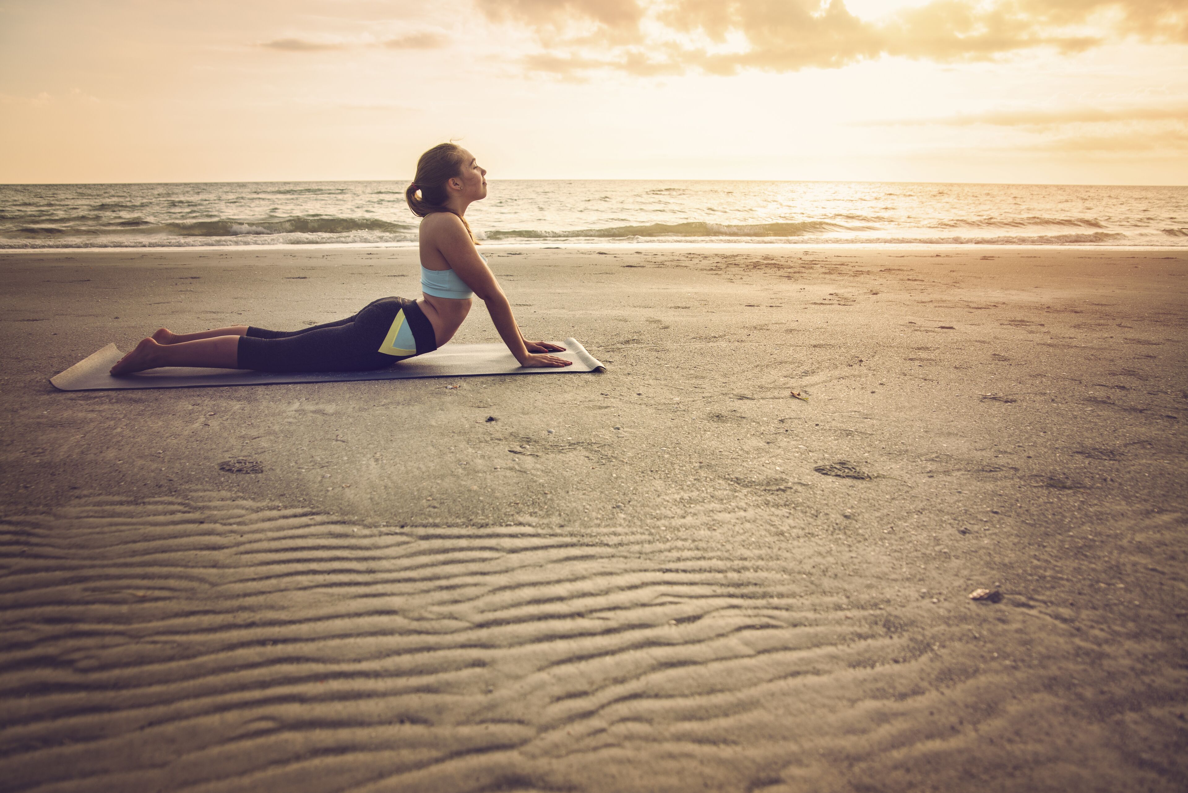 Une personne pratiquant le yoga sur un tapis, sur une plage, avec l’océan en arrière-plan.