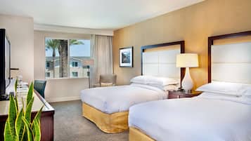 A hotel room with two beds, a desk, a chair, a lamp, and a view of palm trees outside.
