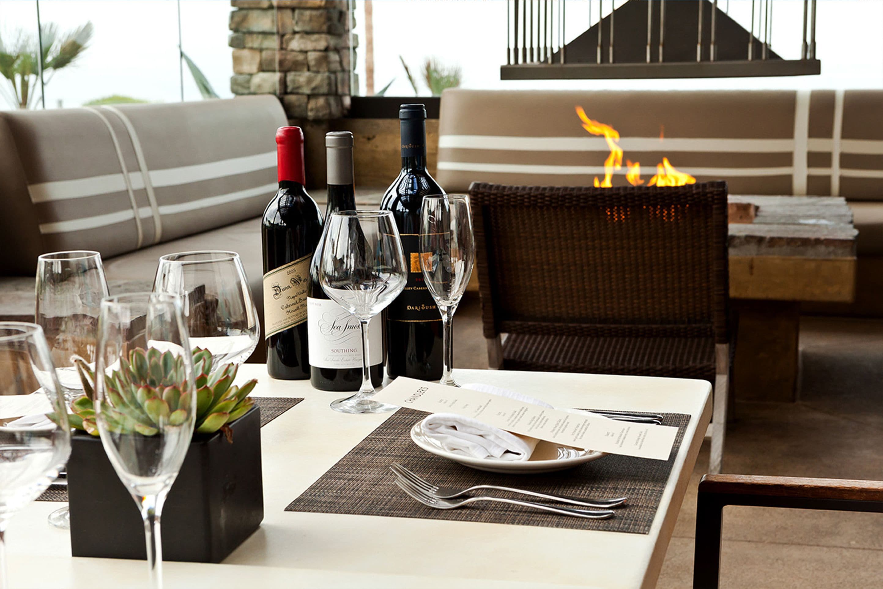 A dining table set with wine bottles, glasses, and a menu, with a fire feature in the background.