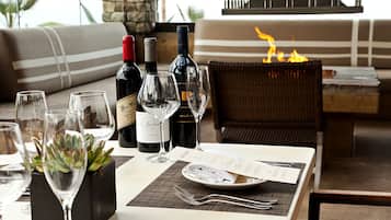 A dining table set with wine bottles, glasses, and a menu, with a fire feature in the background.