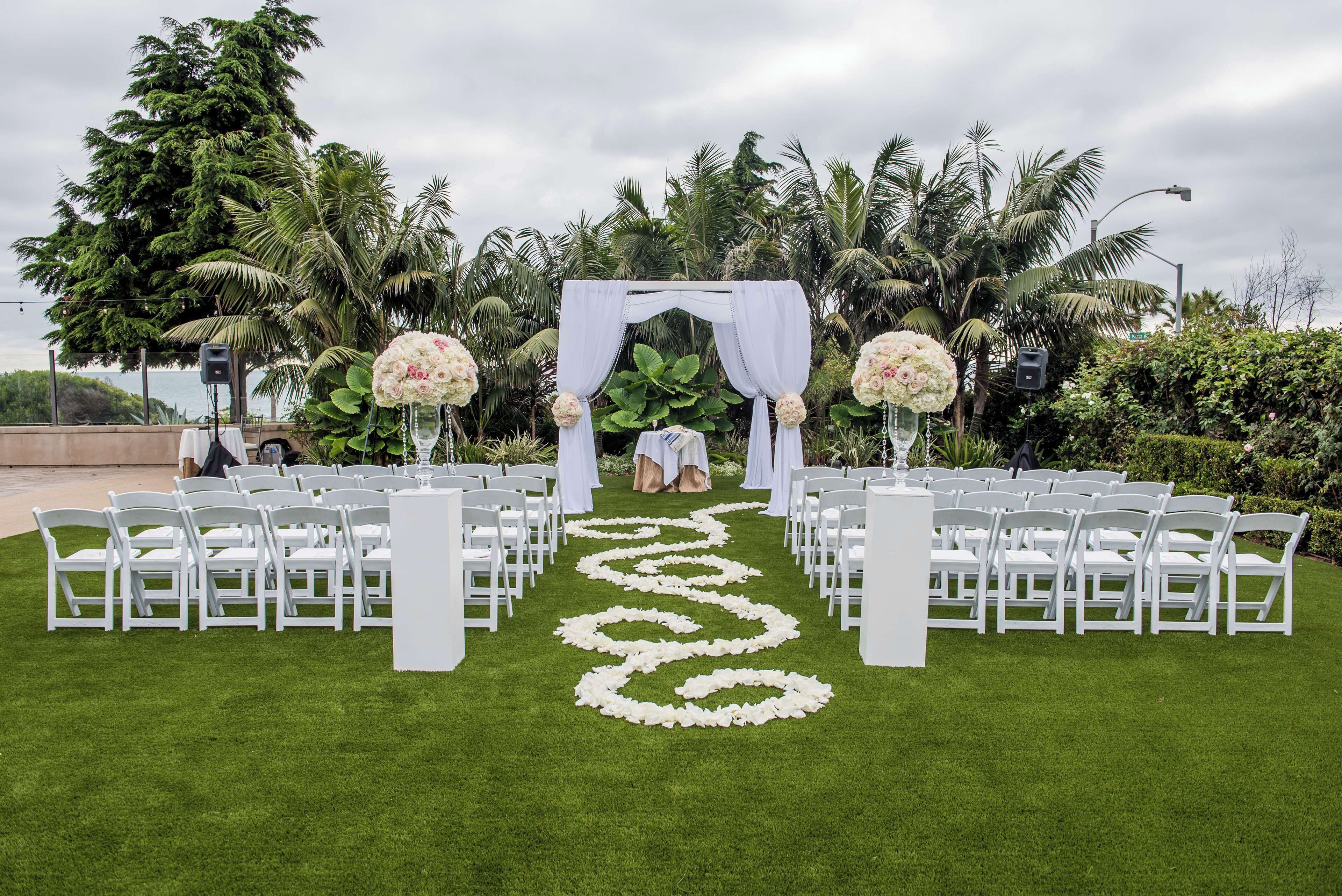 A wedding ceremony setup with a white aisle, floral arrangements, and guests seated on white chairs.