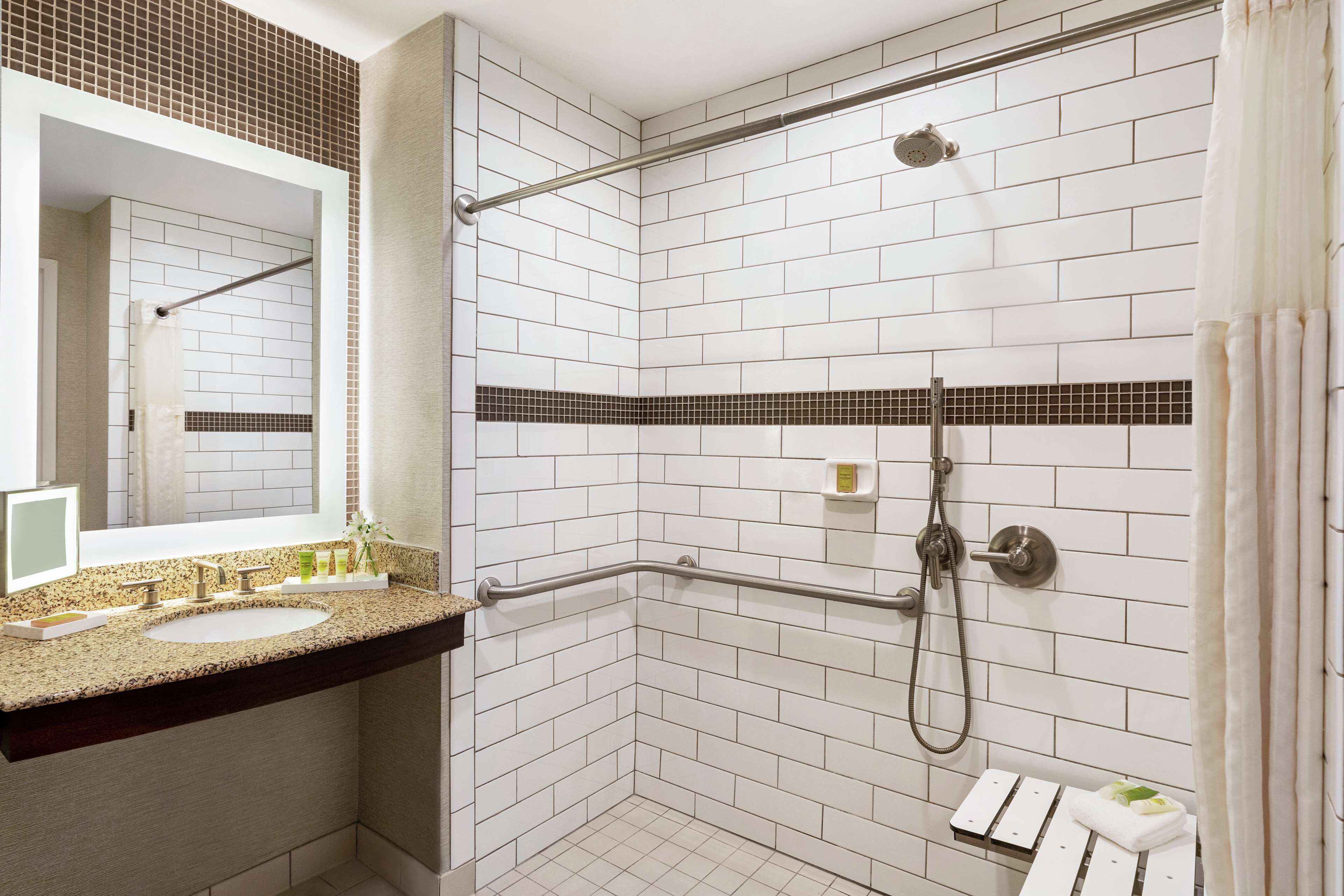 Ein Badezimmer mit weißen Fliesen, einer Granit-Arbeitsplatte, einem Waschbecken, einem Spiegel, einer Dusche mit Duschbrause und einem Handtuchhalter.