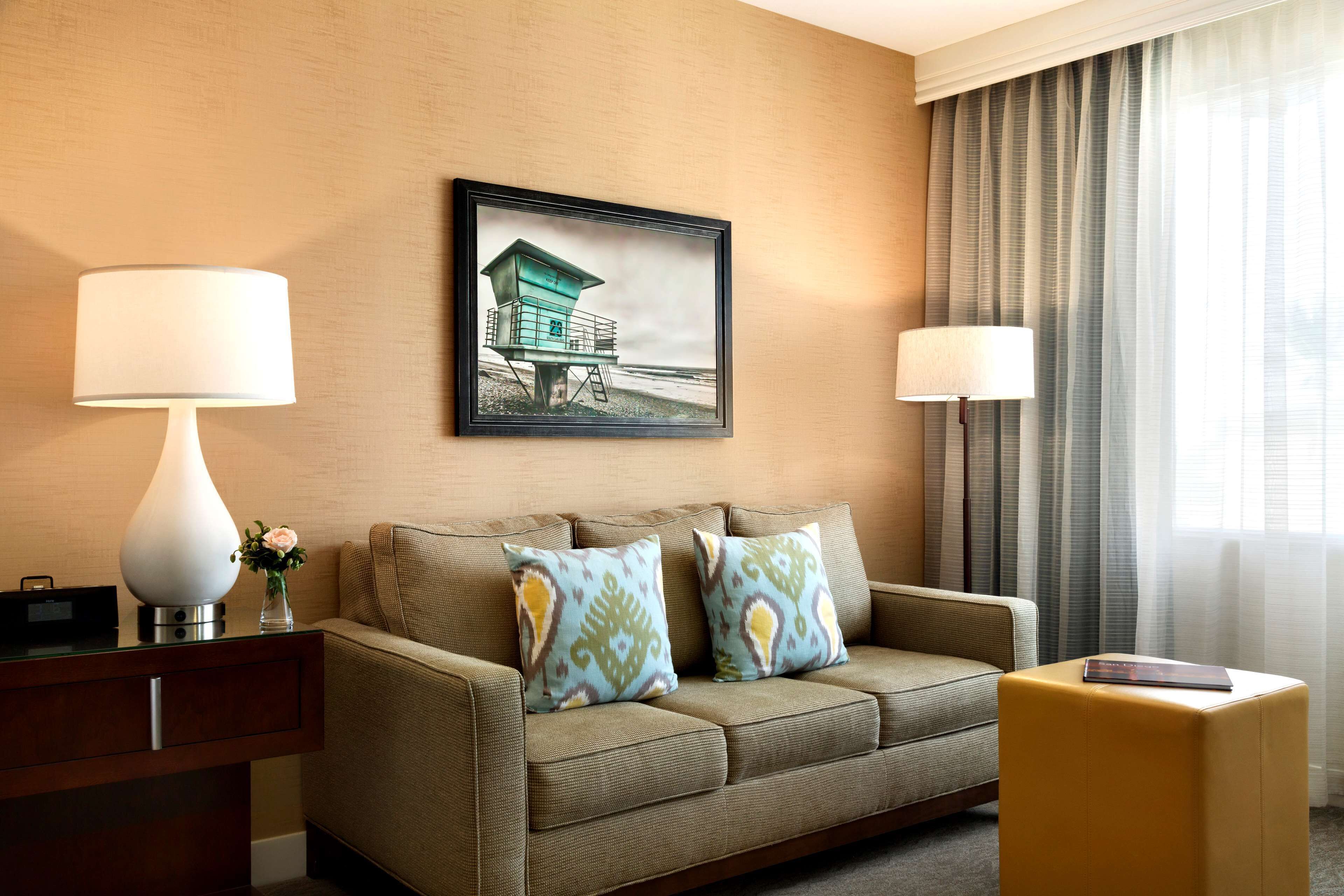 A hotel room with a beige sofa, two patterned throw pillows, a framed picture of a lifeguard tower, a bedside table with a lamp, and a small bedside table with a book.