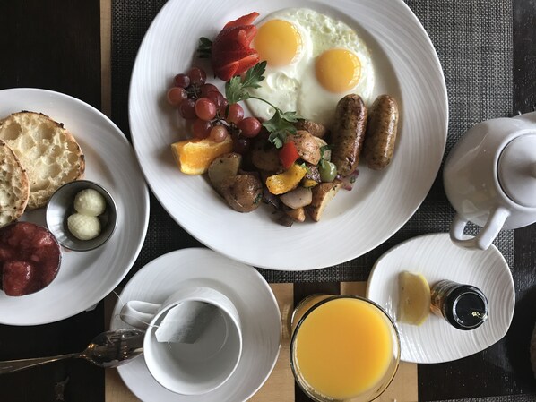 A breakfast setting with plates of eggs, sausages, toast, and fruit, along with a cup of coffee and a glass of orange juice.