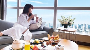 Una mujer sentada en un sofá junto a una ventana con vistas a la ciudad, disfrutando de una comida con varios platos y bebidas.