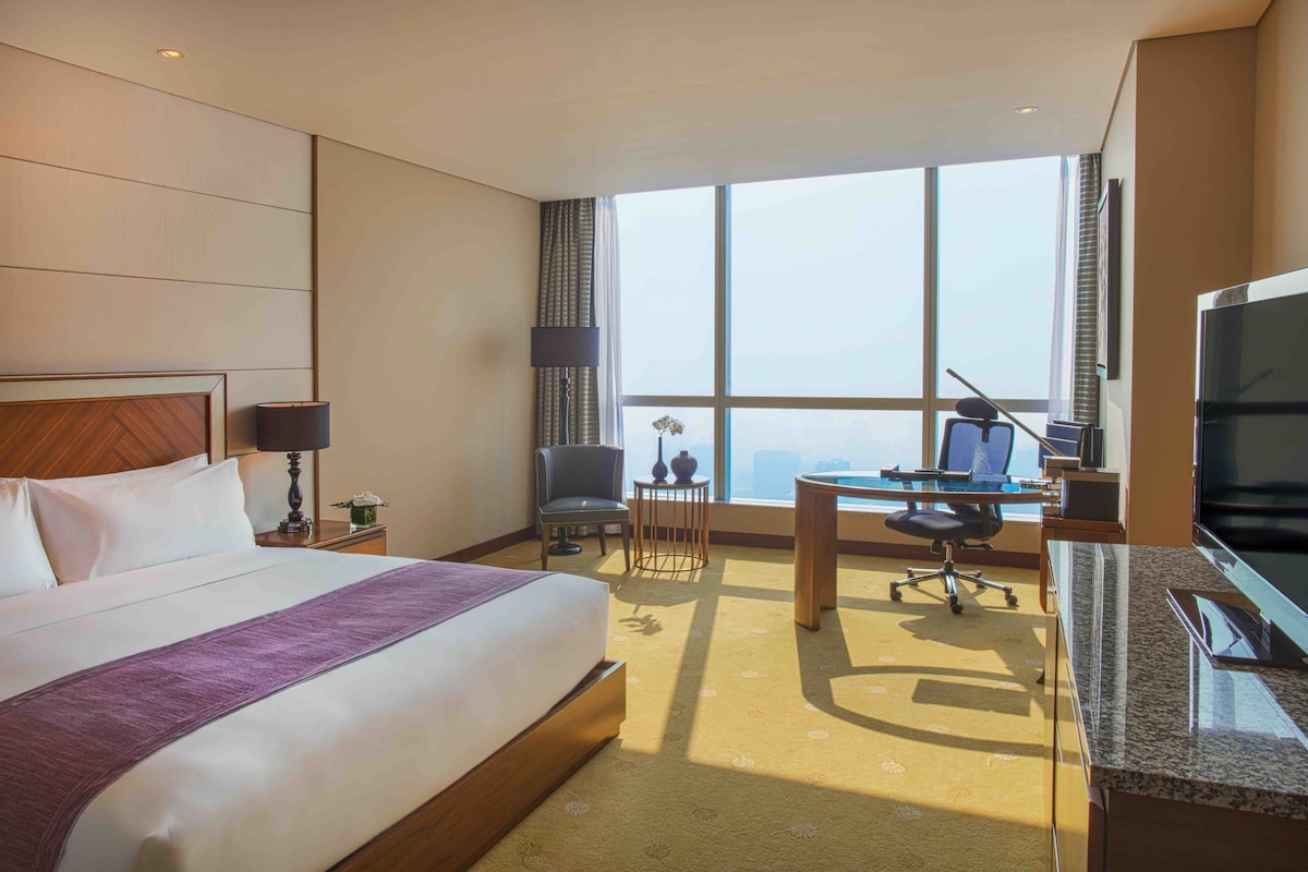Een hotelkamer met een groot bed, een bureau, een stoel, een televisie en uitzicht op de stad.