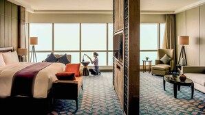 Habitación de hotel con una cama grande, un sofá, un escritorio y una silla. Se ve un ventanal con cortinas, una lámpara de pie y un televisor montado en la pared.
