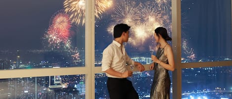 Una pareja de pie junto a un amplio ventanal con vistas a un paisaje urbano y un espectáculo de fuegos artificiales.