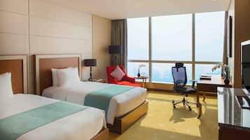 A hotel room with two beds, a desk, a chair, a TV, and a large window with a city view.