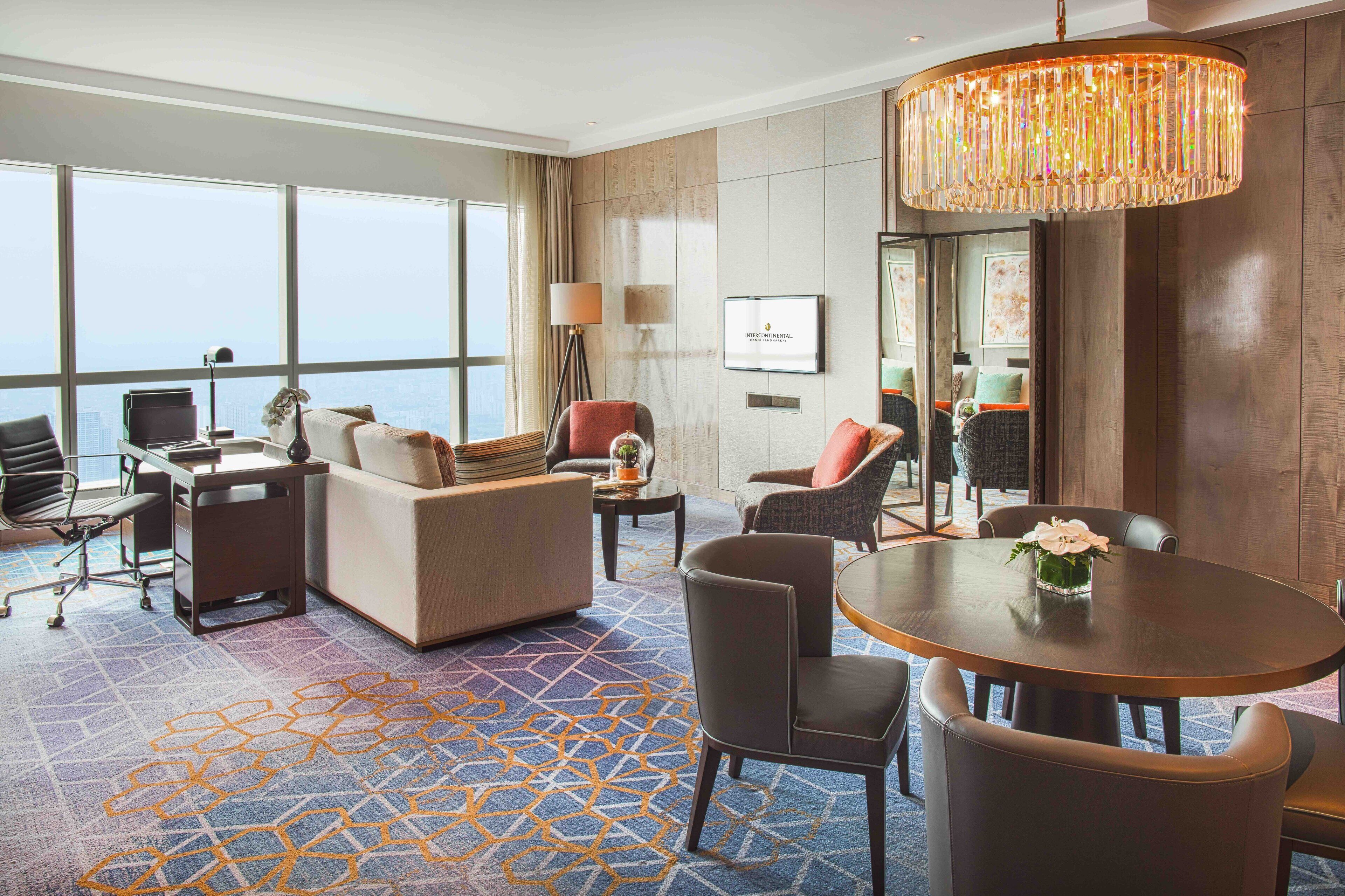 A hotel room with a large window, a round dining table, a sofa, a desk with a computer, and a chandelier.