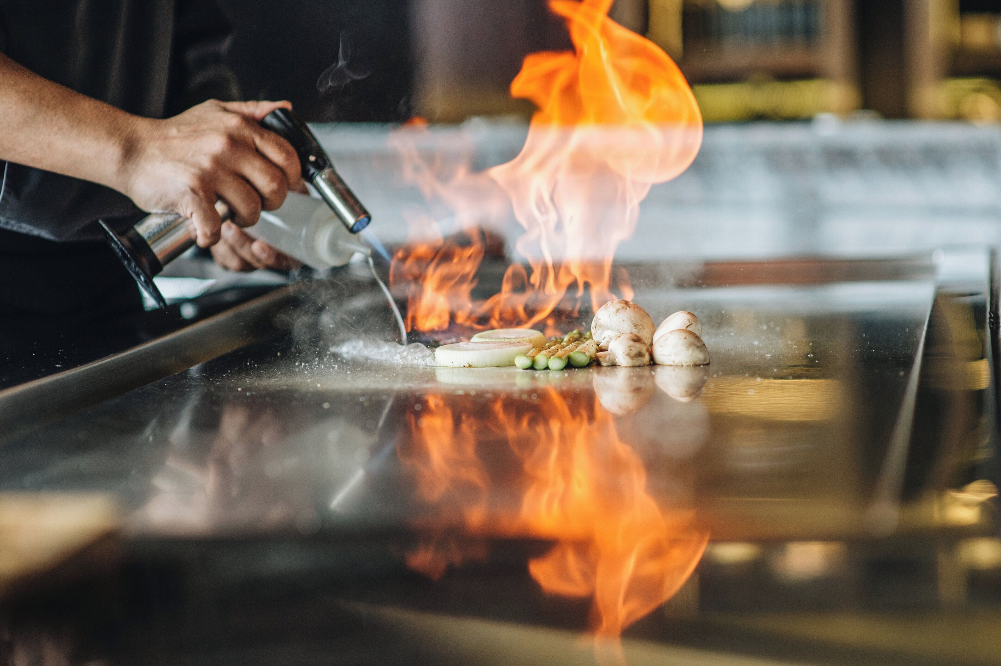 A person cooking on a hot griddle with vegetables and meat, flames visible above the griddle.