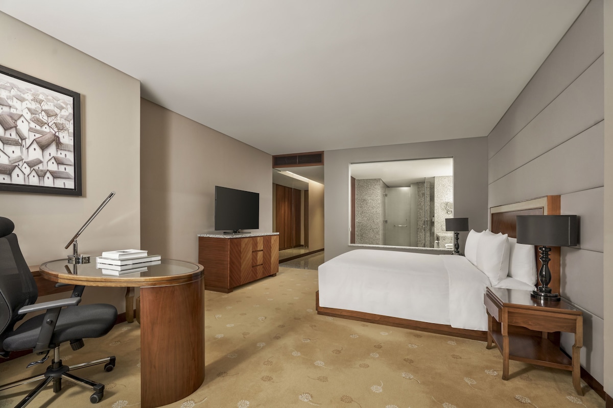 Hotelkamer met een bed, een bureau met stoel, een televisie, een spiegel en een zichtbare badkamer door een geopende deur.