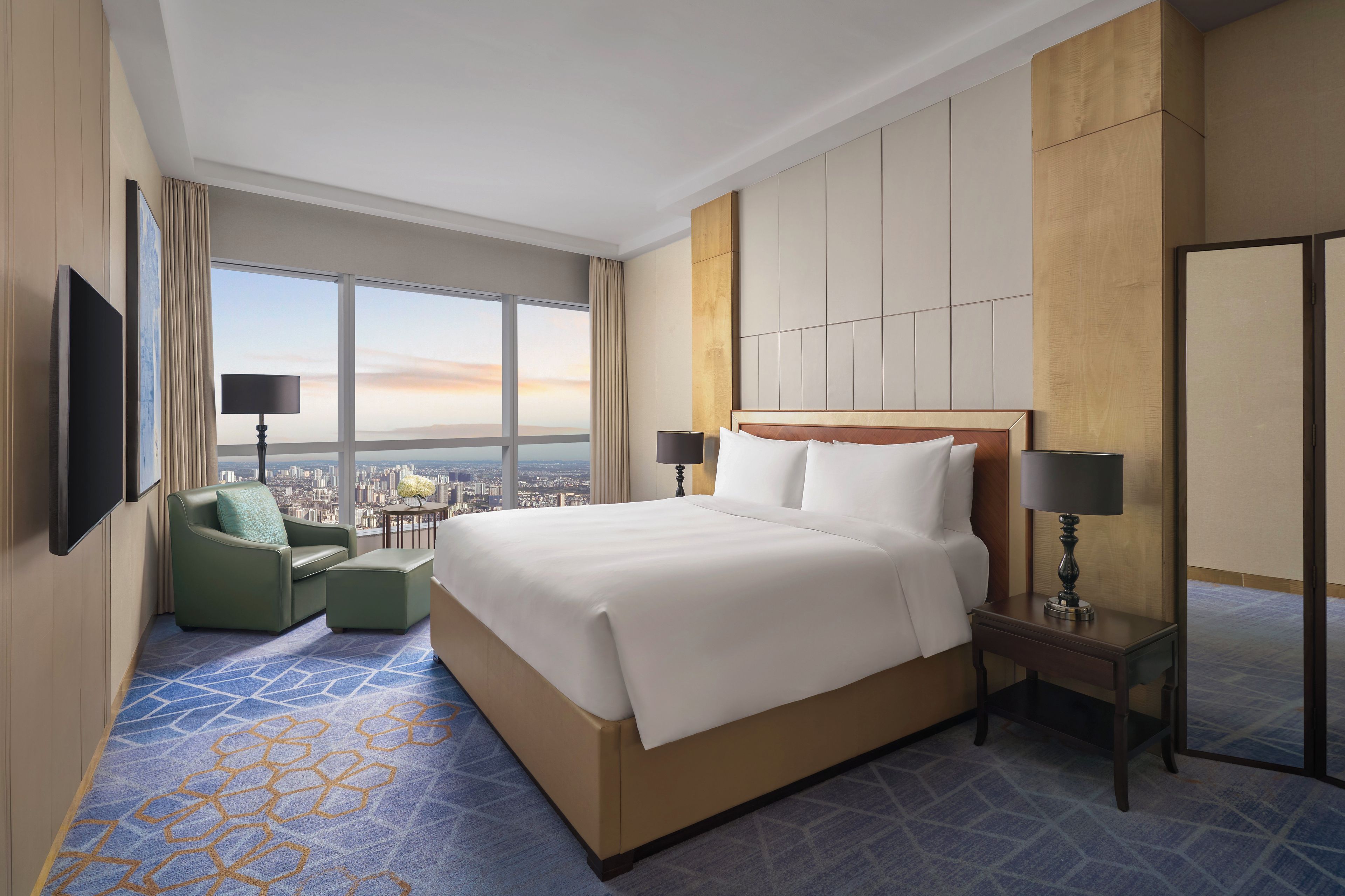 A hotel room with a large bed, a green armchair, a nightstand with a lamp, and a view of the cityscape through large windows.