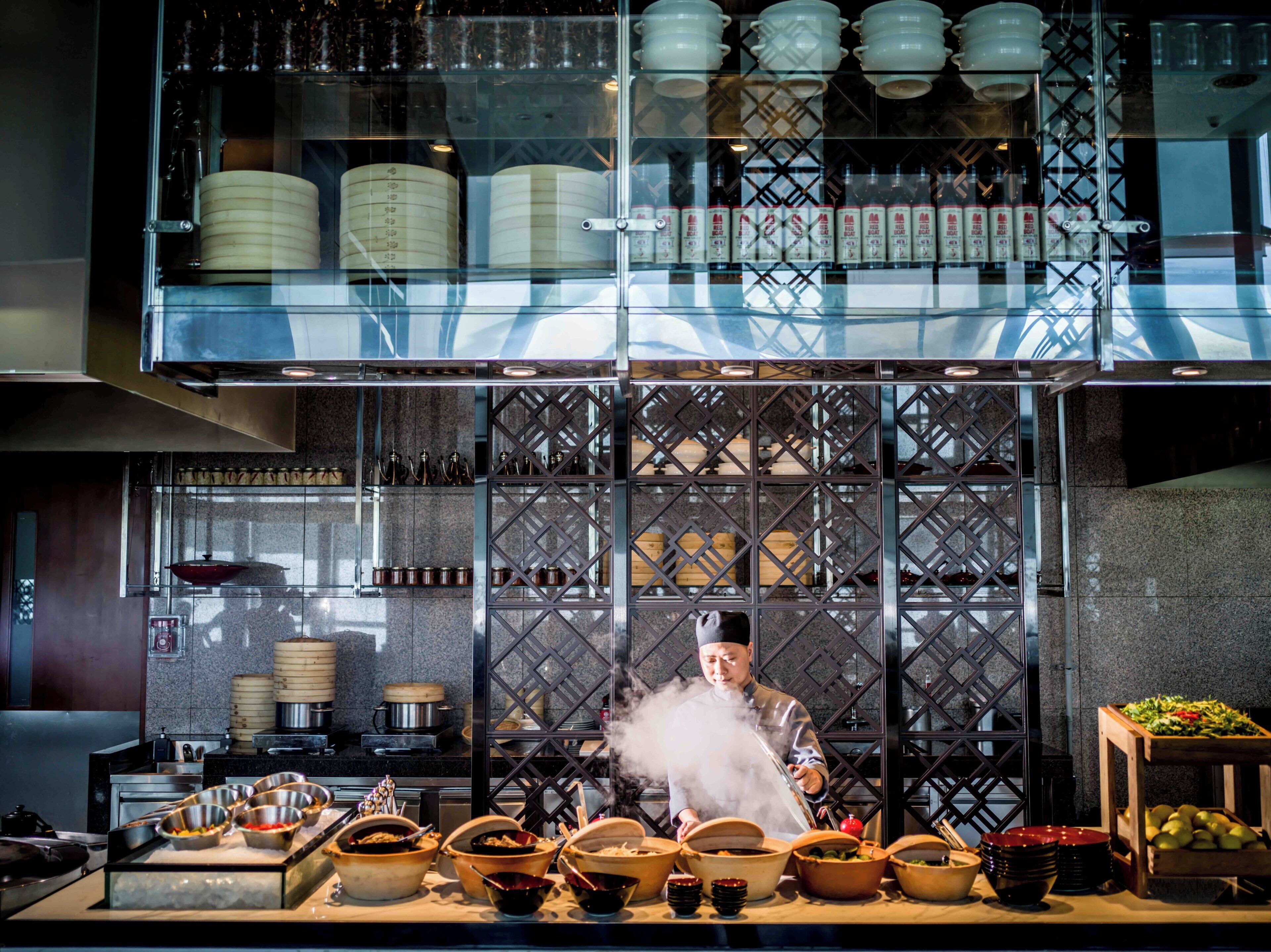 Un chef prépare un plat dans une cuisine moderne, où la vapeur s'élève, et où une variété d'ustensiles de cuisine et d'assiettes sont disposés sur le plan de travail.