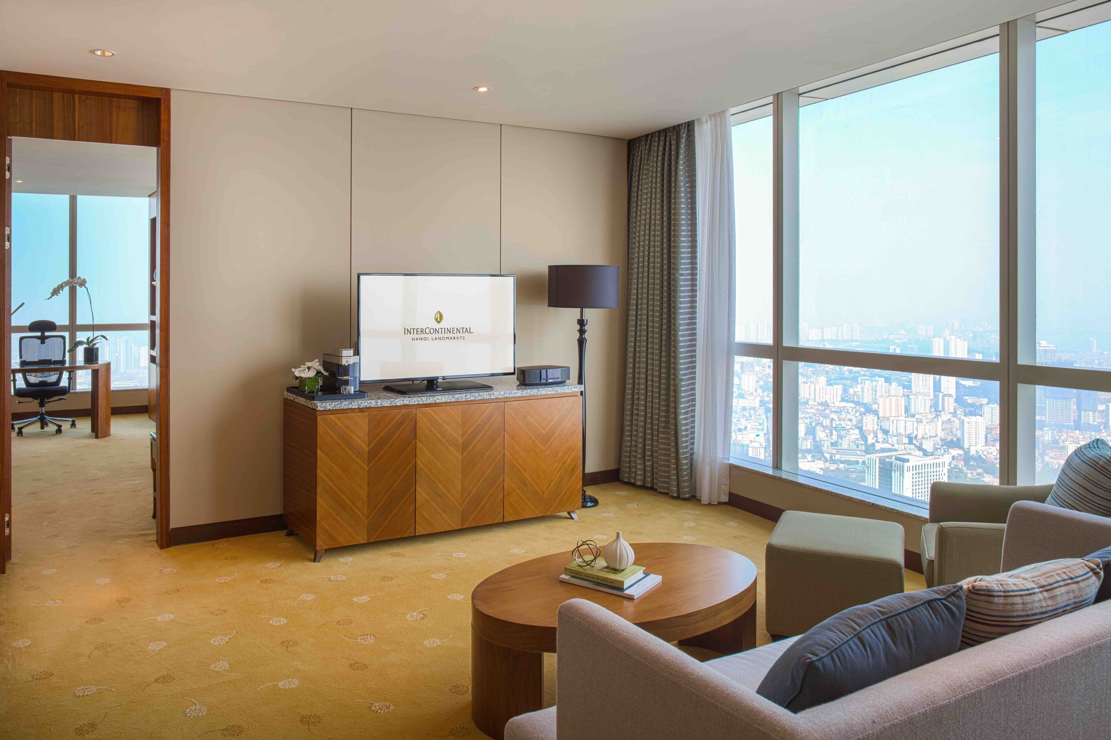 Une chambre d’hôtel avec une grande fenêtre, un téléviseur posé sur un meuble en bois, une table basse ronde et un canapé.