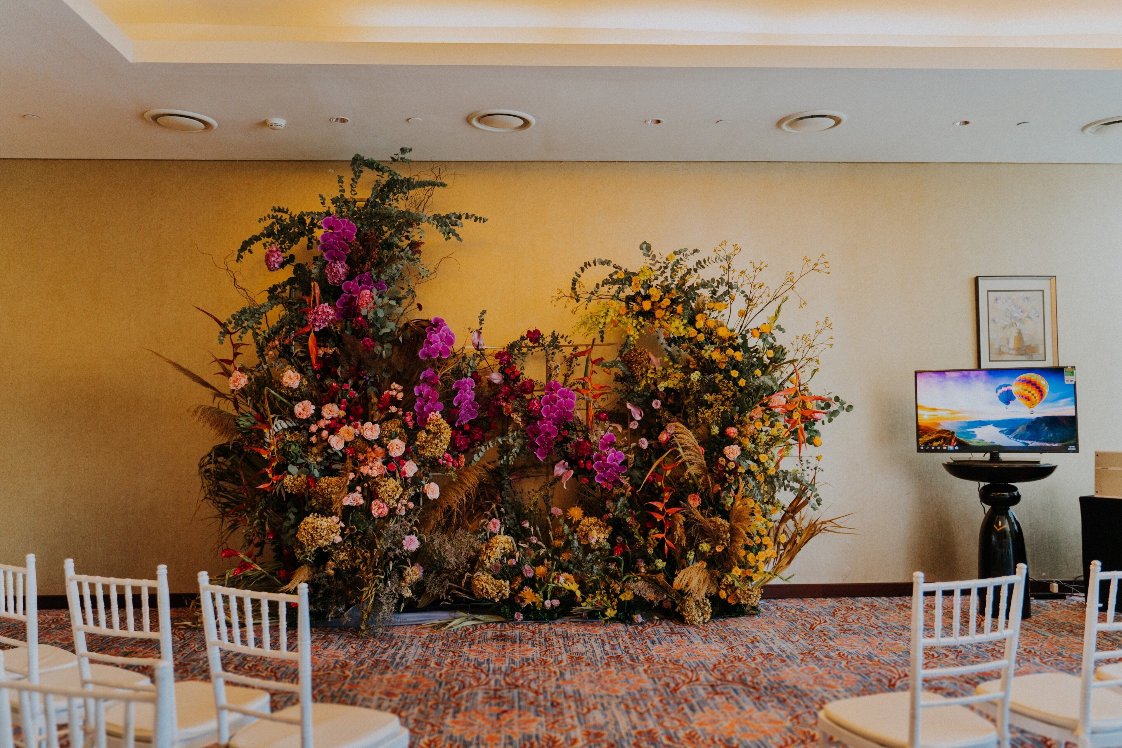 A room with a floral arrangement, white chairs, a carpeted floor, and a television on a stand.