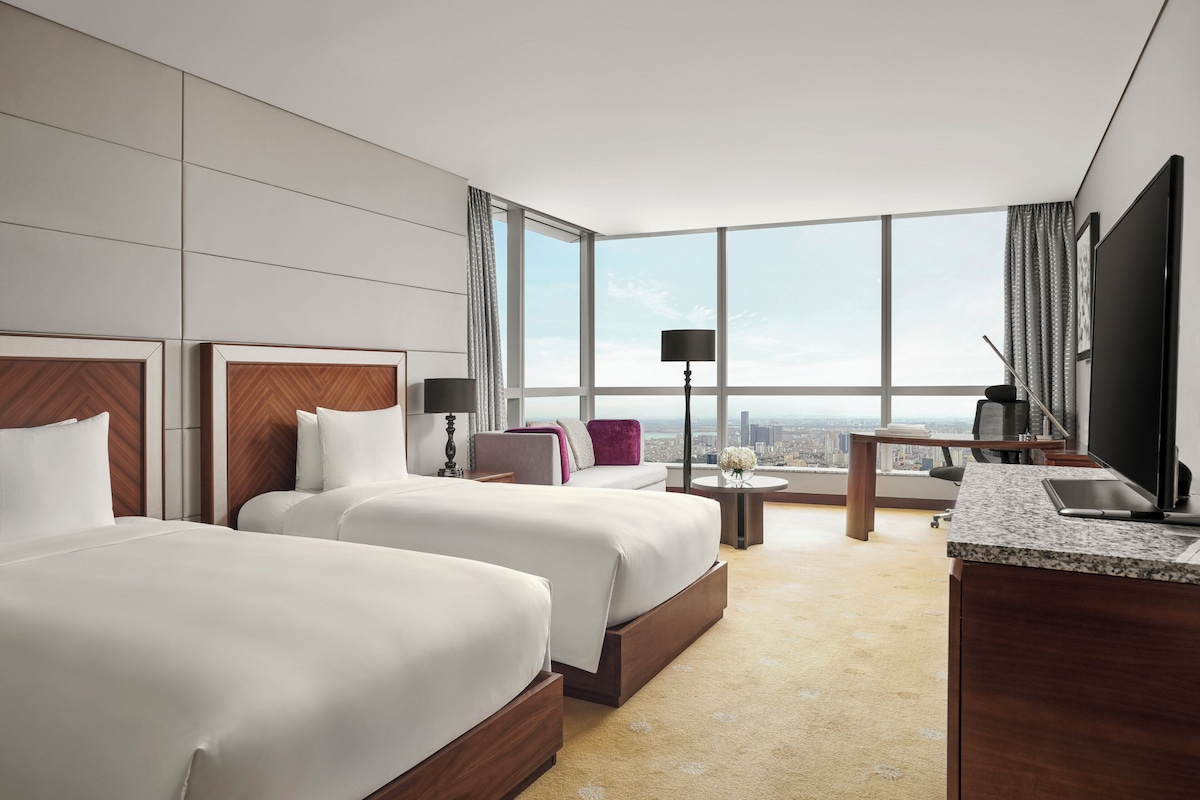 Een hotelkamer met twee bedden, een bureau, een stoel, een televisie en een groot raam met uitzicht op de stad.
