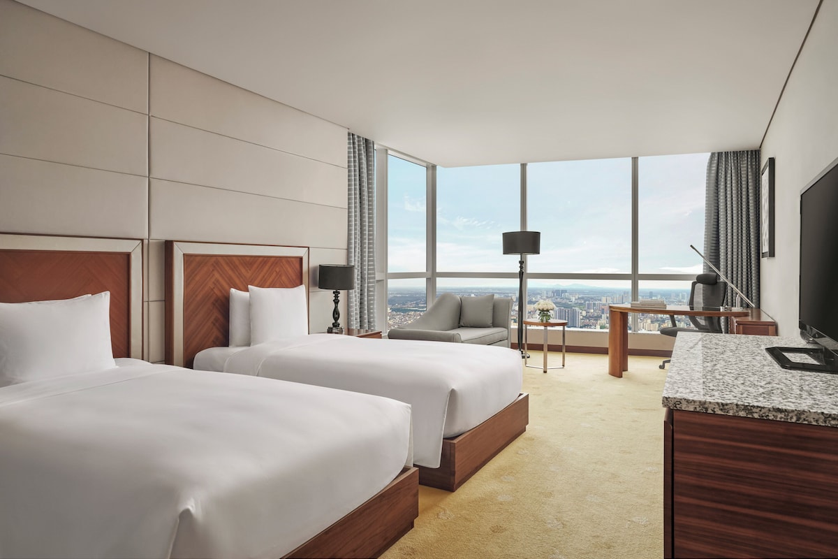 Een hotelkamer met twee bedden, een bureau, een stoel, een televisie en uitzicht op de stad.