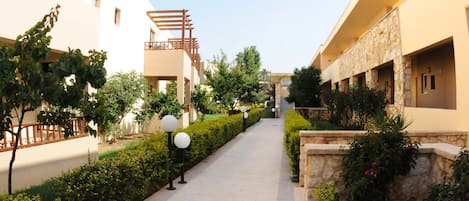 A pathway flanked by neatly trimmed hedges and trees, leading to a building with a stone facade and a balcony.