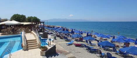 A pool area with lounge chairs and umbrellas, a beach with sun loungers and umbrellas, and a view of the sea and mountains.