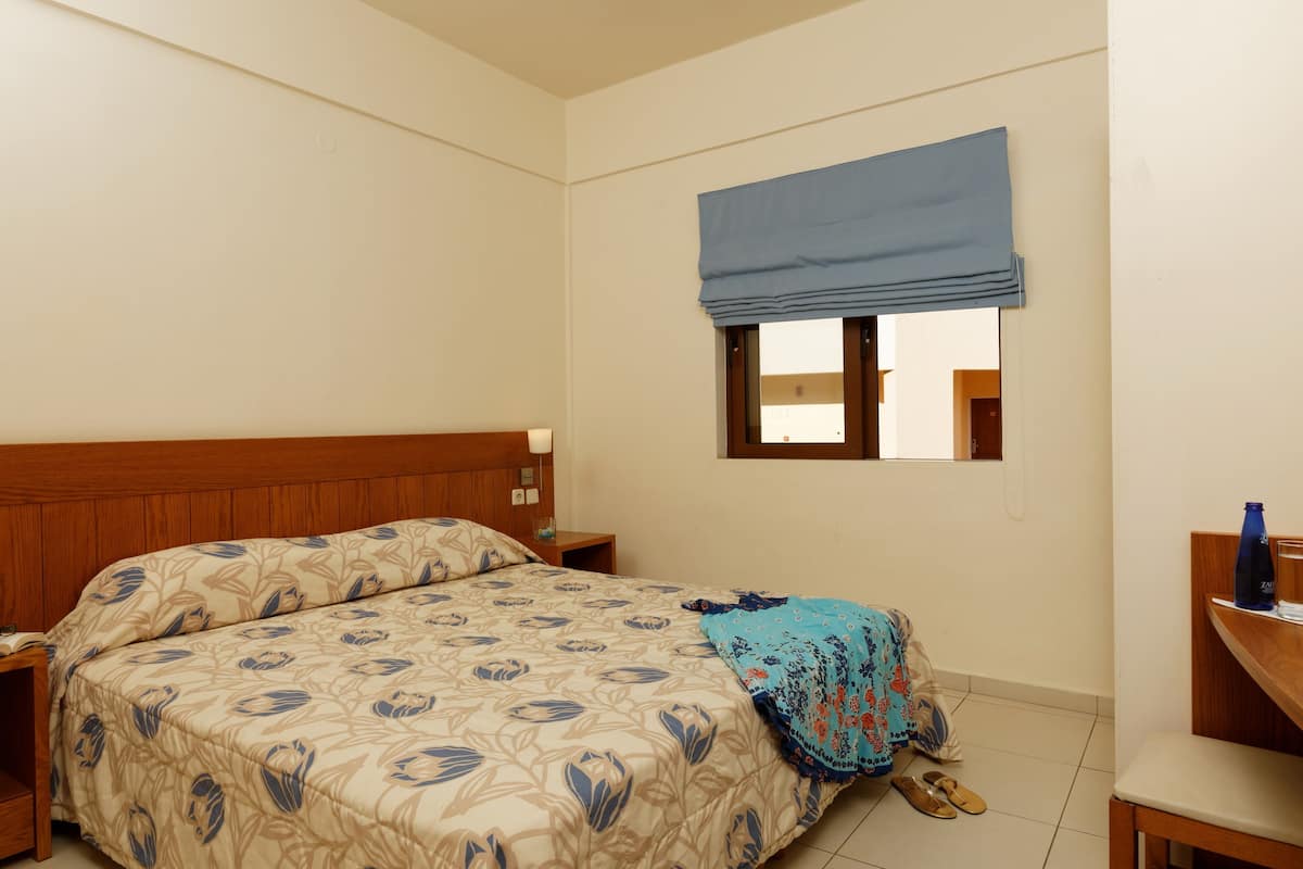 Une chambre avec une tête de lit en bois, un lit recouvert d’une couvre-lit à motifs floraux, un lambrequin bleu à la fenêtre, une table de chevet avec une lampe et une chaise sur laquelle est posée une bouteille bleue.