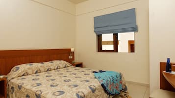 Un dormitorio con cabecera de madera, una cama con colcha de estampado floral, un visillo azul en la ventana, una mesita de noche con una lámpara y una silla con una botella azul encima.