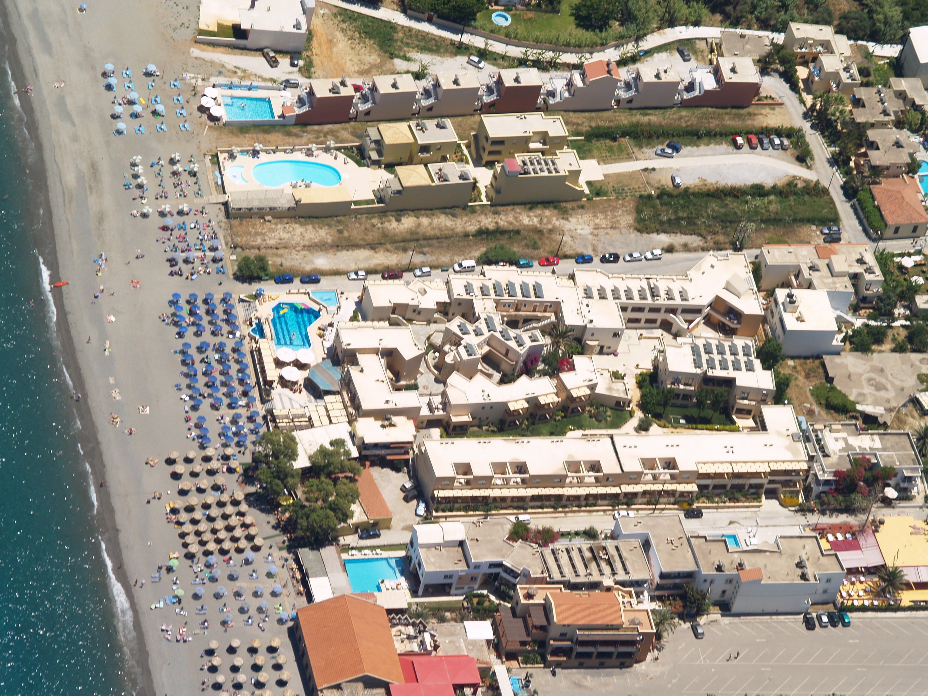 An aerial view of a coastal resort with multiple buildings, a beach with umbrellas, and a swimming pool.