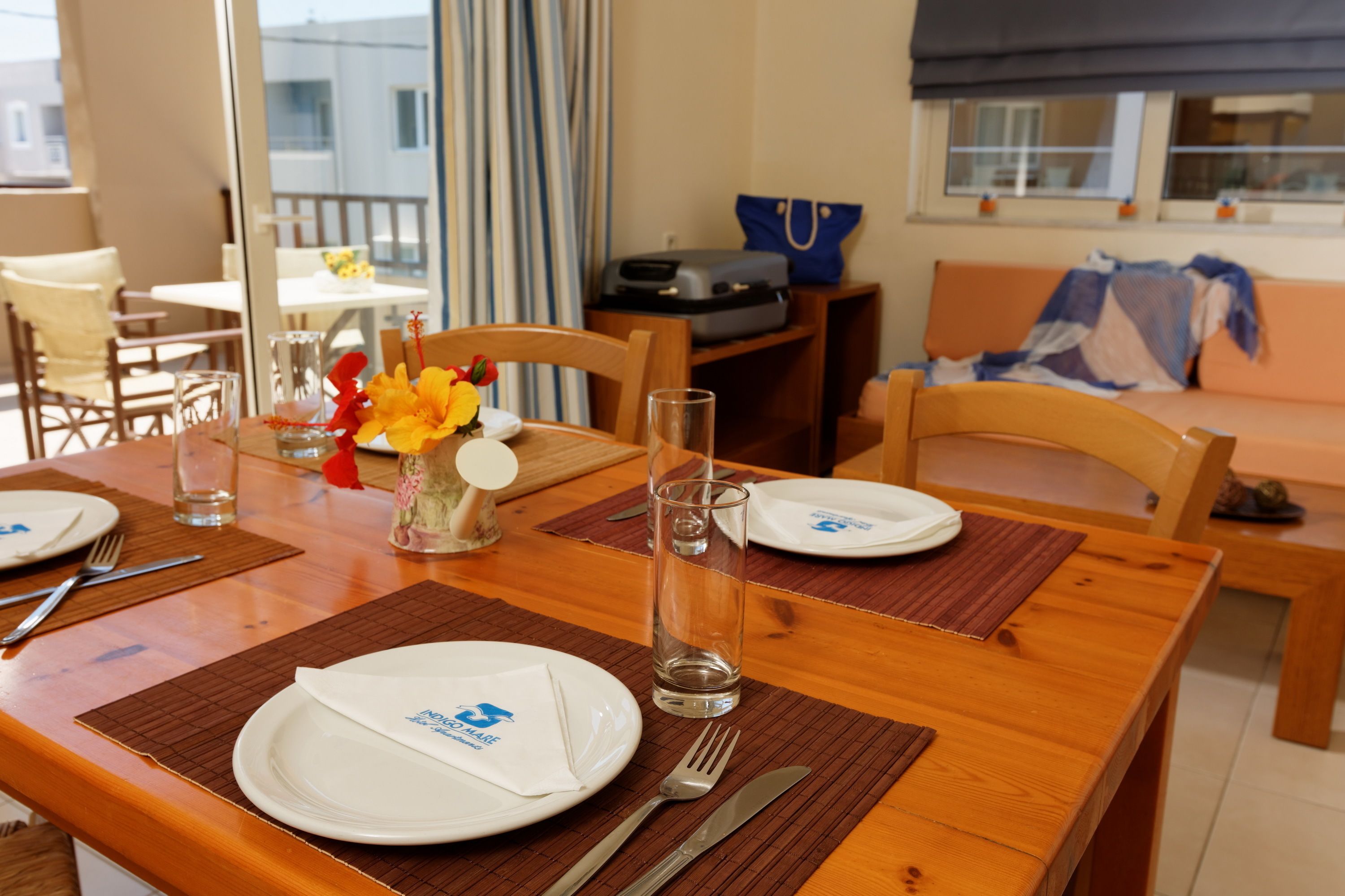 Mesa de comedor preparada con platos, cubiertos y vasos, con vista al balcón y un sofá al fondo.