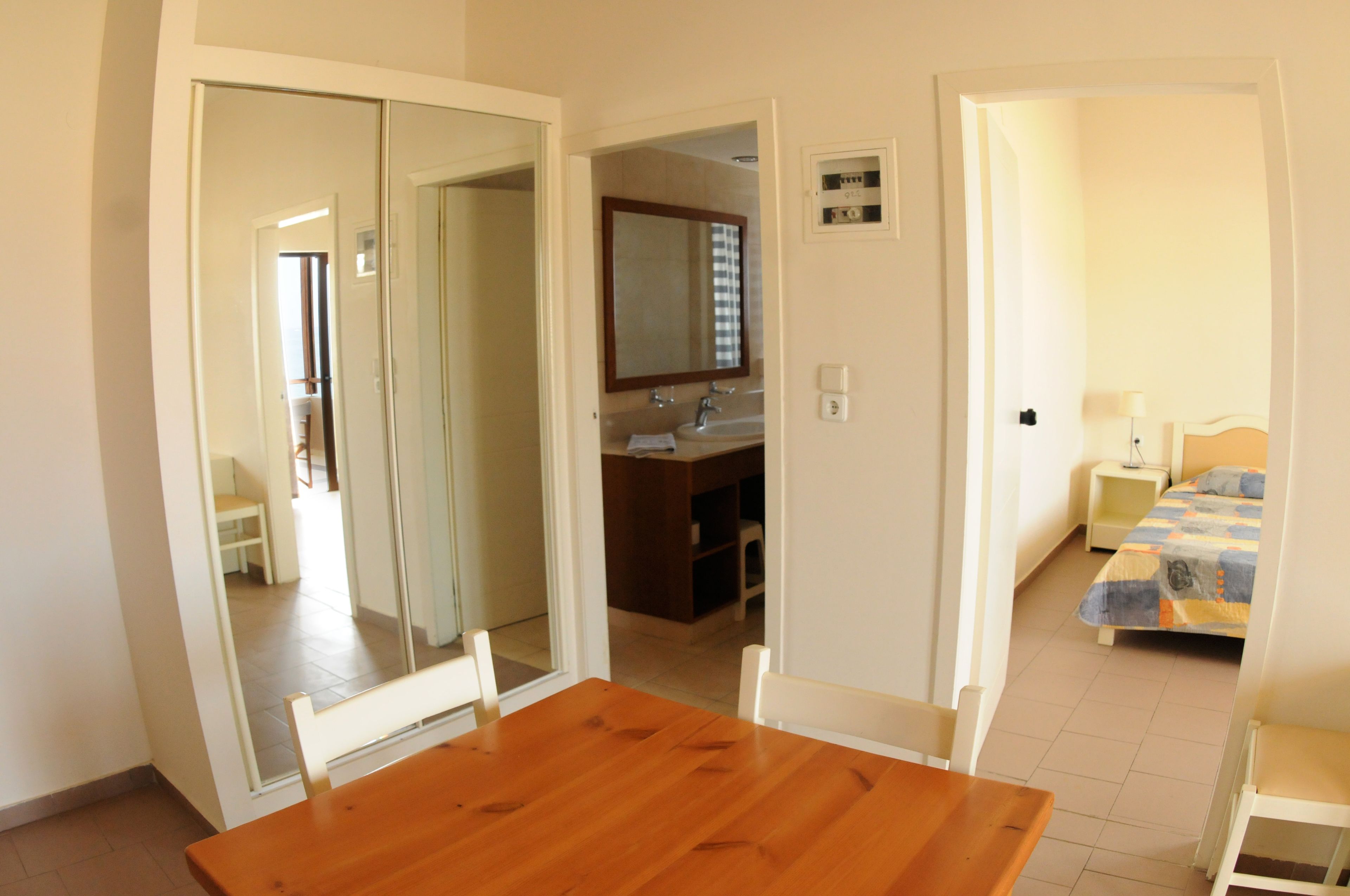 A room with a wooden dining table, a mirror, a sink, a bed, and a nightstand.