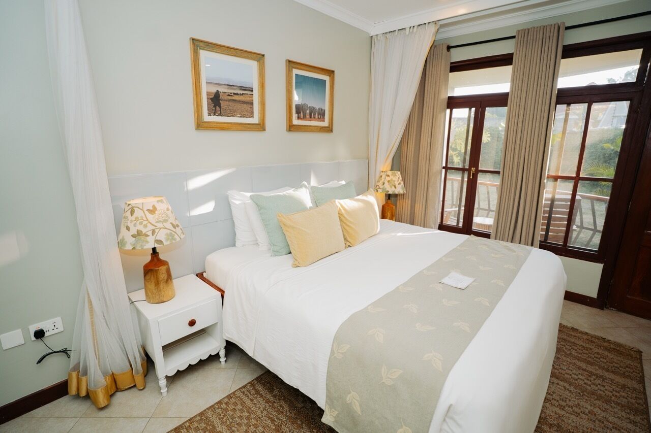 A hotel room with a large bed, two framed pictures on the wall, a nightstand with a lamp, and a view through a glass door.