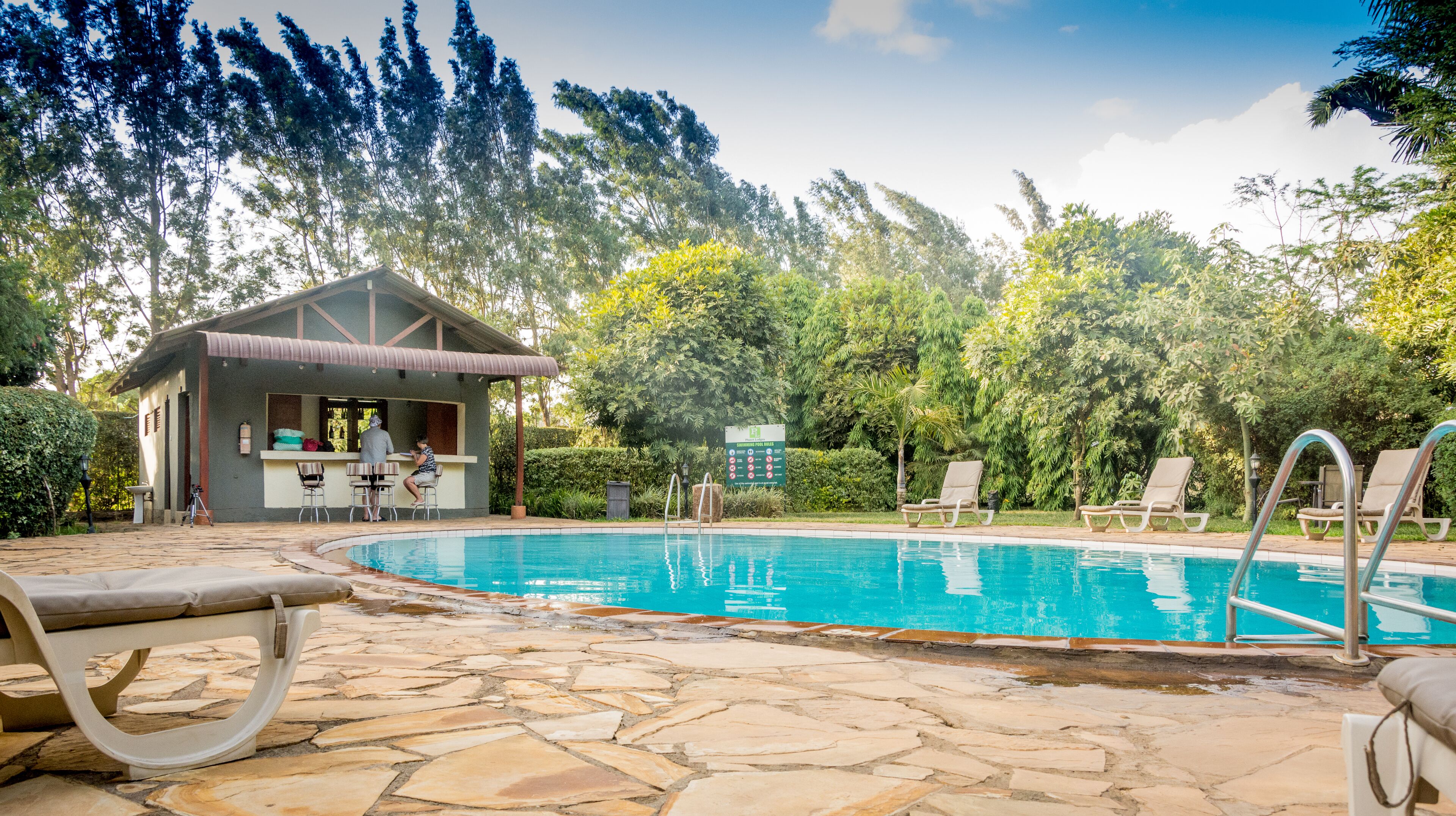 A pool area with lounge chairs, a bar, and a small building with a roof.