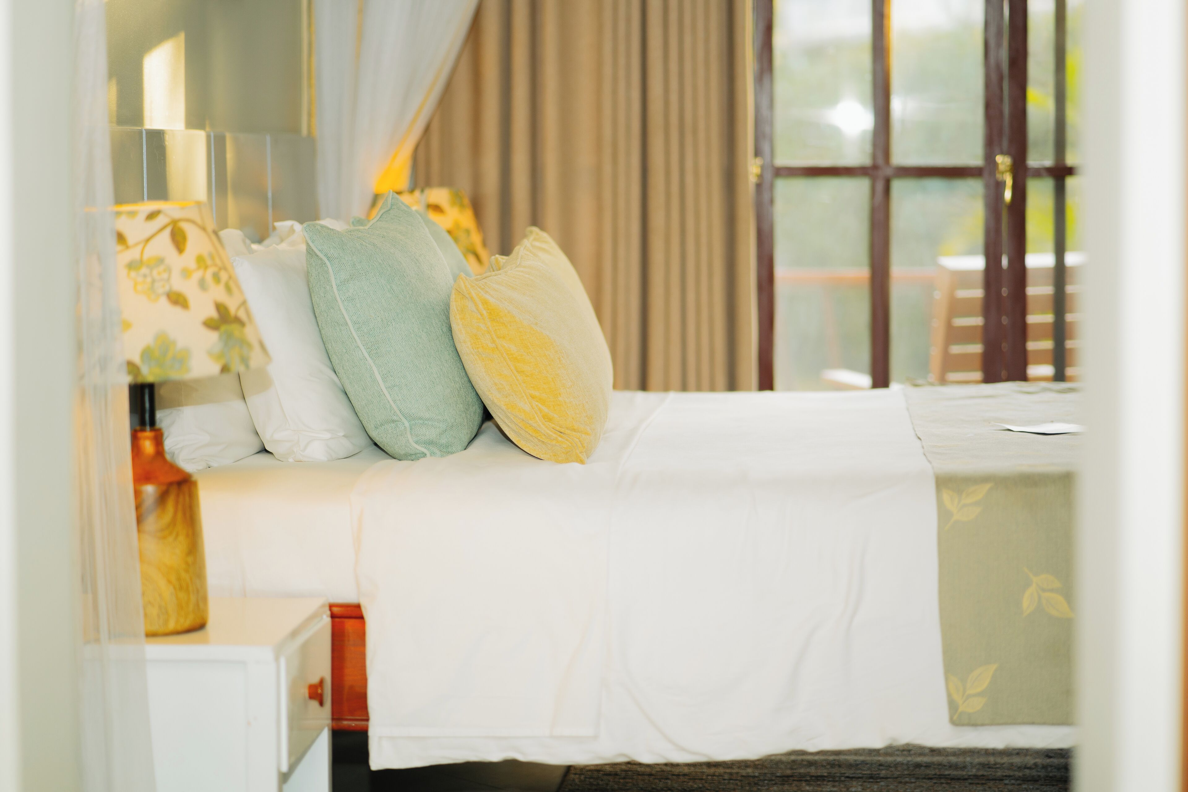 Una cama bien hecha con ropa blanca y almohadas decorativas, una mesita de noche con una lámpara y una ventana con cortinas.