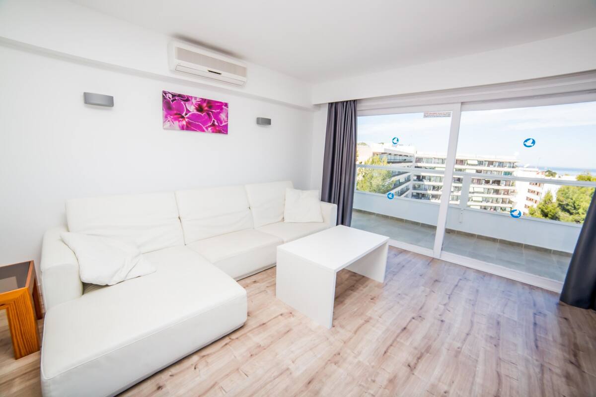 Een moderne woonkamer met een witte bank, een klein witte tafeltje en een grote glazen schuifdeur die leidt naar een balkon met uitzicht op de stad.