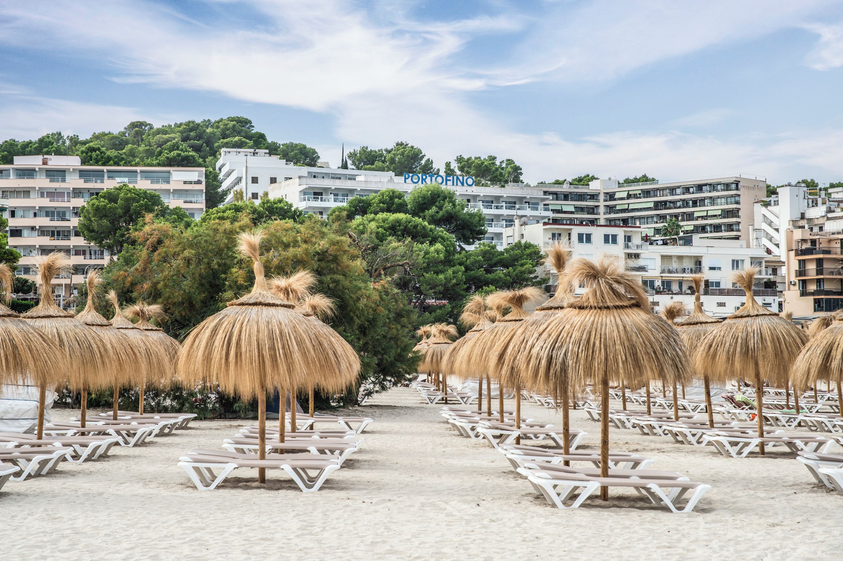 Ein Strand mit Liegestühlen und Reetdachschirmen, ein klarer Himmel und ein Gebäude im Hintergrund mit der Aufschrift 'PORTOFINO'.